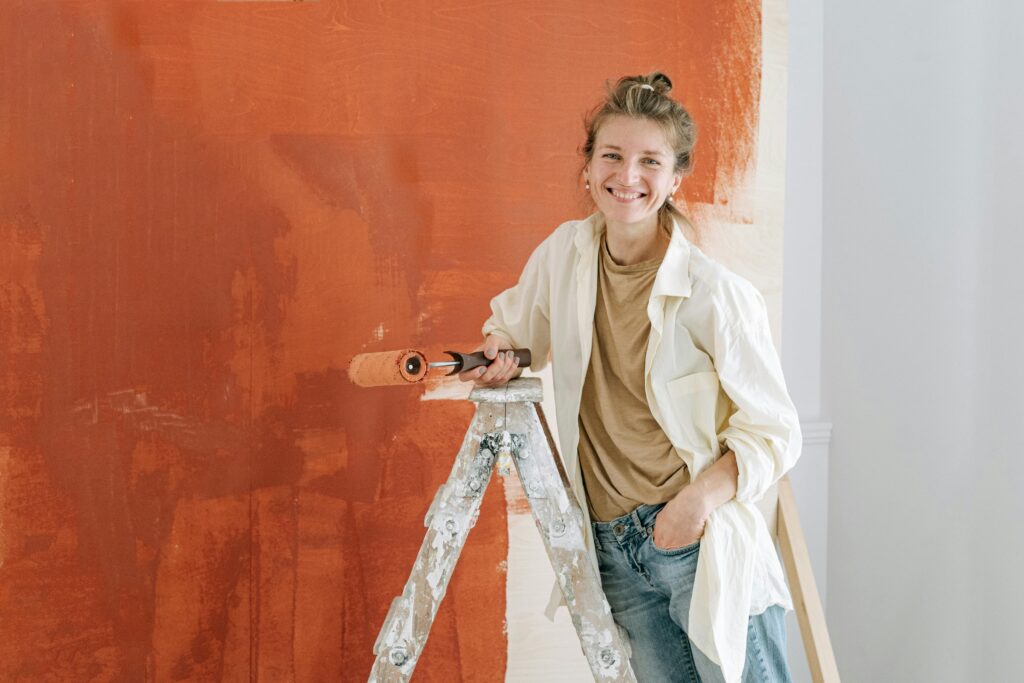 A cheerful woman in casual attire paints an orange wall while standing on a ladder indoors.