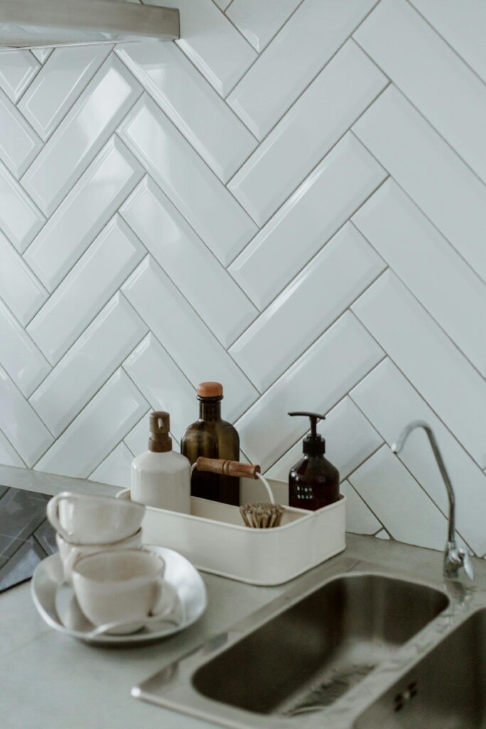 Stylish kitchen sink area with cleaning products and cups on a tiled counter.